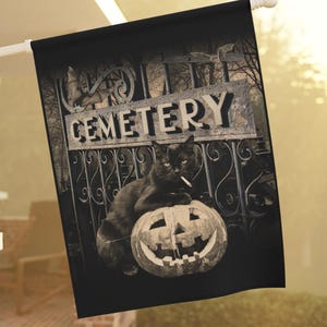 Könnte beinhalten: Schwarze Flagge mit einer schwarzen Katze, die eine Zigarette raucht und auf einem geschnitzten Kürbis sitzt. Die Flagge zeigt ein Friedhofstor-Design mit dem Wort "CEMETERY". Eine Halloween-Dekoration.