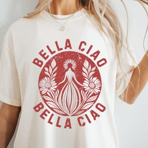 以下が含まれることがあります： クリーム色のTシャツ。赤い円形のグラフィックデザインで、「BELLA CIAO」の文字と、花に囲まれた人物のイラストが中央に描かれています。ヴィンテージ風のデザインです。