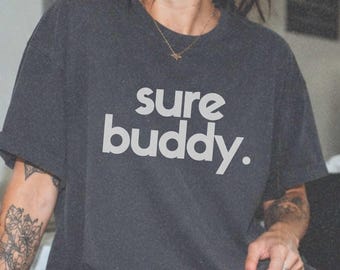 May include: A charcoal gray short-sleeve t-shirt featuring the text "sure buddy." printed in a light gray, rounded sans-serif font across the chest. The garment has a crew neckline and a relaxed fit, constructed from a soft, cotton-like fabric.