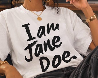 May include: A white short-sleeved t-shirt featuring the black text I am Jane Doe. printed in a bold, handwritten style across the chest. The garment has a crew neckline and a relaxed fit, constructed from a plain white fabric with black lettering.