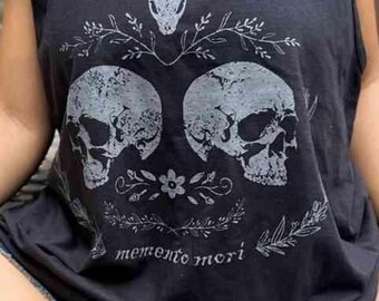 Puede incluir: Camiseta sin mangas negra con un gráfico de dos calaveras y detalles florales. El texto "memento mori" está impreso debajo. La camiseta se combina con pantalones cortos vaqueros.