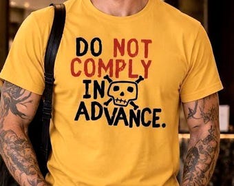 May include: A mustard yellow short-sleeved t-shirt featuring the text DO NOT COMPLY IN ADVANCE in red and black lettering, accompanied by a black and white skull and crossbones graphic centered on the chest of the garment.