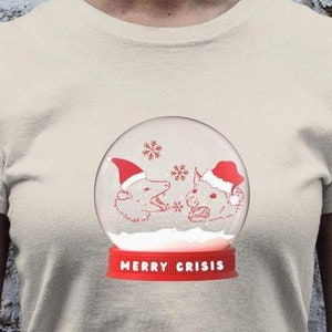 Peut inclure: T-shirt crème avec un motif de boule à neige. À l'intérieur, deux animaux de dessin animé portant des bonnets de Père Noël. La base de la boule à neige porte l'inscription "MERRY CRISIS" en rouge. Des flocons de neige tombent.