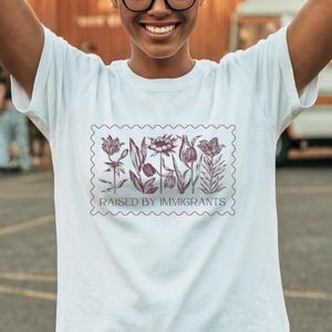 Peut inclure: T-shirt blanc avec un motif de timbre floral et le texte "RAISED BY IMMIGRANTS". Le t-shirt est porté par une personne portant des lunettes. L'arrière-plan comprend une caravane et des guirlandes lumineuses.