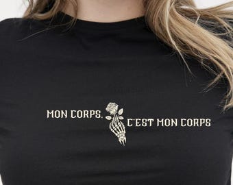 May include: A black short-sleeved t-shirt featuring white text that reads MON CORPS, C'EST MON CORPS. Between the two phrases is a graphic illustration of a skeletal hand holding a single rose, rendered in white and light gray tones.