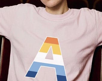 May include: A light pink t-shirt featuring a large, colorful letter "A" in orange, yellow, white, and blue. The person is sitting in a theater seat with red upholstery. The shirt is short-sleeved.