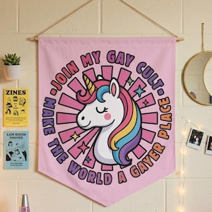 Może przedstawiać: Różowy baner z grafiką jednorożca i napisem "Join My Gay Cult, Make the World a Gayer Place" wisi na białej ceglanej ścianie. Pokój jest ozdobiony lampkami choinkowymi, tęczową kołdrą i pluszowymi jednorożcami.
