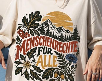 May include: An off-white t-shirt featuring a graphic print of mountains, a sun, flowers, and acorns. The text "MENSCHENRECHTE FÜR ALLE" is printed in the center in red and yellow. The shirt has short sleeves and a crew neckline.