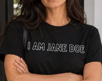 May include: A black short-sleeved t-shirt featuring the white outlined text I AM JANE DOE printed across the chest. The shirt is paired with dark charcoal denim jeans, a black leather belt with a gold triangular buckle, and a black shoulder bag.