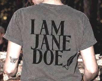 May include: A charcoal gray short-sleeve t-shirt featuring the black text I AM JANE DOE printed on the back, accompanied by a small black quill pen graphic. The shirt has a crew neckline and a standard fit, worn with dark denim jeans.