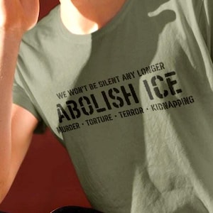 May include: Olive green t-shirt with the text "WE WON'T BE SILENT ANY LONGER ABOLISH ICE" and the words "MURDER - TORTURE - TERROR - KIDNAPPING" in black. The person is holding a skateboard.