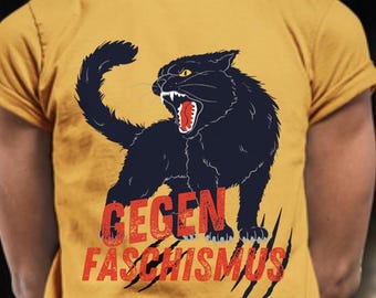 May include: A mustard yellow short-sleeved t-shirt featuring a graphic of a snarling black cat with yellow eyes and an open mouth. Below the cat, the red distressed text reads "GEGEN FASCHISMUS" over black claw marks on the fabric.