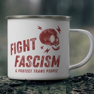May include: White enamel mug with a silver rim and a white handle. The mug features the words "FIGHT FASCISM & PROTECT TRANS PEOPLE" in red, along with a red skull and lightning bolt graphic.
