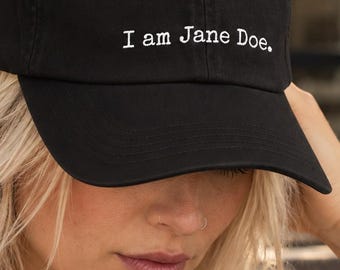 May include: A black baseball cap featuring the white embroidered text "I am Jane Doe." on the front panel. The cap has a structured crown, a curved brim, and a button on top, providing a classic look with clear, legible white lettering.