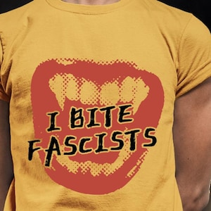 Può includere: T-shirt giallo senape con grafica rossa di una bocca con zanne e la scritta nera "I BITE FASCISTS". La maglietta ha maniche arrotolate e girocollo.