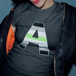 Könnte beinhalten: Eine Person trägt ein dunkelgraues T-Shirt mit einem großen schwarzen Buchstaben 'A' auf der Vorderseite. Der Buchstabe 'A' ist mit den Farben der asexuellen Pride-Flagge gefüllt, die schwarz, grau, weiß, grün und hellgrün sind. Die Person trägt auch eine schwarze Lederjacke.