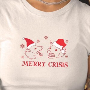 Peut inclure: T-shirt blanc avec un motif rouge de deux opossums portant des bonnets de Père Noël et le texte "MERRY CRISIS". Le design comprend des flocons de neige. Vêtement décontracté et festif.