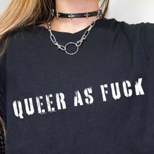 Puede incluir: Camiseta negra con la frase "QUEER AS FUCK" en blanco y con letras desgastadas. La camiseta se combina con un collar choker negro y una manga de red.
