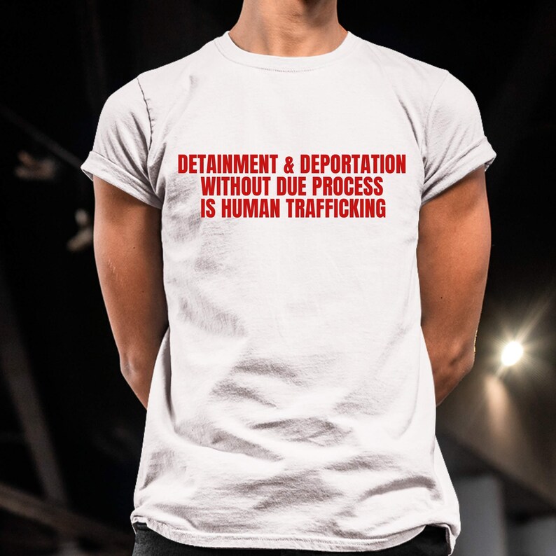 May include: White t-shirt with the red text: DETENTION & DEPORTATION WITHOUT DUE PROCESS IS HUMAN TRAFFICKING. The shirt has short sleeves and a crew neck.