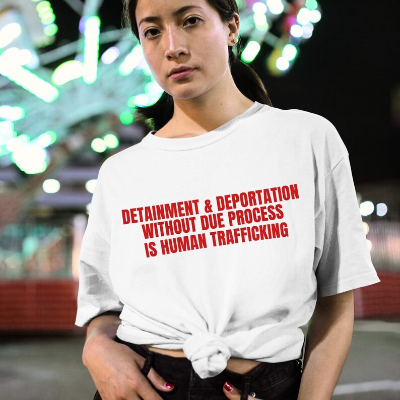 May include: White t-shirt with the red text: DETAINMENT & DEPORTATION WITHOUT DUE PROCESS IS HUMAN TRAFFICKING. The shirt is knotted at the waist. The background is blurred with colorful lights.