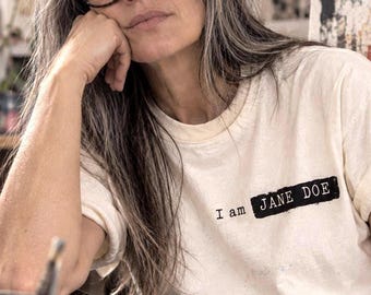 May include: A cream-colored short-sleeve t-shirt featuring the black text "I am JANE DOE" printed on the chest. The text "JANE DOE" is highlighted by a solid black rectangular block background, while the shirt has a crew neckline.