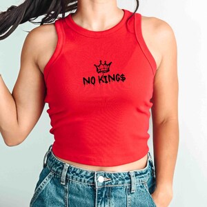 May include: A red ribbed tank top with the text "NO KINGS" printed in black, along with a crown graphic. The top is sleeveless and has a round neckline. The model is wearing blue denim jeans.