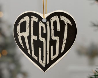 May include: A heart-shaped ceramic ornament featuring a black background with the word RESIST printed in large, distressed white capital letters. The ornament is suspended by a thin gold-colored string attached to a small metal loop at the top.