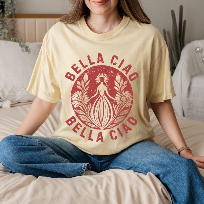 以下が含まれることがあります： 赤色の「BELLA CIAO」の文字が、赤い花の模様を囲むようにプリントされた、薄い黄色のTシャツ。青いジーンズと合わせて着用されています。デザインは、中央の人物と花の要素が特徴です。