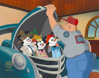 May include: Animation cel featuring the Warner Bros. characters Yakko, Wakko, and Dot in a car with a mechanic. The car is blue, and the mechanic wears a red cap and gray shirt. The Warner Bros. logo is visible in the lower left corner.