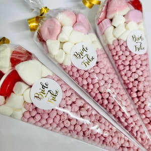 May include: Three clear plastic cones filled with pink and white sweets. Each cone has a white circle sticker with the text "Bride Tribe" in gold lettering.