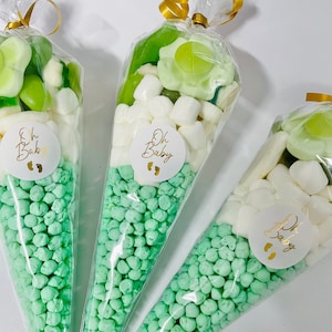 Puede incluir: Tres conos de pl&aacute;stico transparente llenos de caramelos verdes y blancos. Los conos est&aacute;n atados con una cinta dorada y tienen una pegatina circular blanca con el texto "Oh Baby" y un dise&ntilde;o de huella.