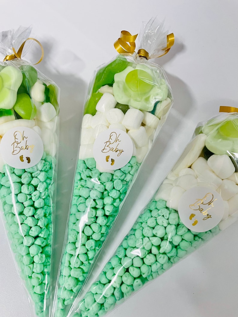 Puede incluir: Tres conos de pl&aacute;stico transparente llenos de caramelos verdes y blancos. Los conos est&aacute;n atados con una cinta dorada y tienen una pegatina circular blanca con el texto "Oh Baby" y un dise&ntilde;o de huella.