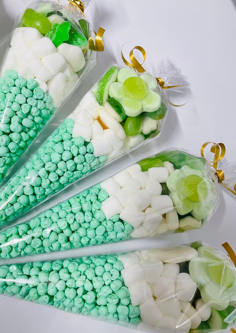 Puede incluir: Cuatro conos de pl&aacute;stico transparente llenos de caramelos verdes y blancos. Los conos est&aacute;n atados con una cinta dorada. Los caramelos incluyen caramelos de gelatina verdes, caramelos gomosos verdes, malvaviscos blancos y granulado verde.