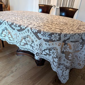 May include: An oval-shaped, off-white lace tablecloth featuring an intricate, repeating floral pattern with openwork mesh detailing. The fabric drapes over the edges of a dark wooden table, showcasing a complex, interconnected design of blossoms and leaves.