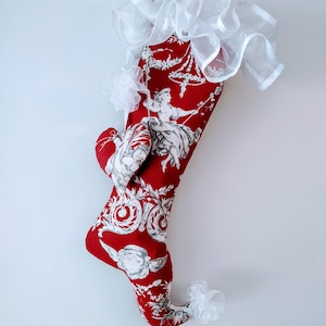 May include: A red Christmas stocking with a white and grey toile pattern featuring a woman and floral designs. The stocking has a white ruffled trim at the top and a white bow accent. A red ribbon loop is attached for hanging.