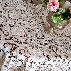Può includere: Una tovaglia in pizzo color bianco sporco caratterizzata da un intricato motivo floreale con viticci e dettagli in rete traforata. Il tessuto presenta un disegno ripetitivo di grandi fiori e foglie, creando una superficie strutturata.