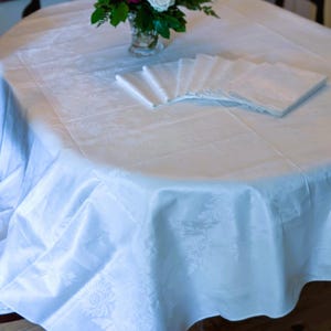 May include: A white, oval tablecloth with a floral design, ready for a meal. A vase of flowers with white, pink, and purple blooms is in the centre. White napkins are neatly arranged on the table, creating an elegant dining scene.
