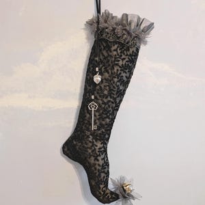 May include: Black lace Christmas stocking with a floral pattern, decorated with silver charms including a key and a heart. The top has a grey ruffled trim. A grey flower and bell decorate the toe. A black ribbon hangs from the top.