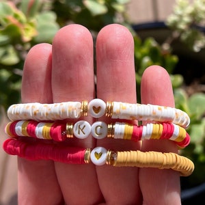 May include: Three colorful beaded bracelets. One is white with yellow dots, another is red and yellow with the letters "KC", and the third is red and yellow. Each bracelet has gold accents and white beads with heart designs.