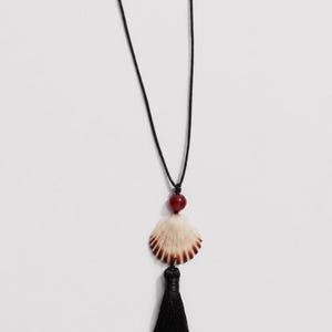 May include: A necklace featuring a long black cord, a single round dark red bead, a fan-shaped seashell with brown and white radial stripes, and a black fabric tassel hanging from the bottom of the shell, creating a layered pendant design.