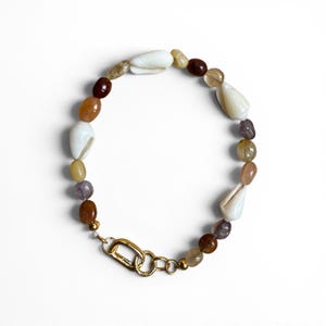 May include: A beaded bracelet featuring a mix of white cowrie shells, translucent yellow, brown, and purple polished stones, and small gold-toned beads. The bracelet is secured with a gold-toned metal lobster clasp and a circular link connector.