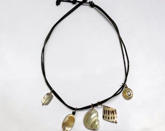 Könnte beinhalten: Eine schwarze Kordelkette mit fünf hängenden Anhängern, darunter ein kleines rechteckiges Muschelstück, eine ovale irisierende Muschel, eine spiralförmige Muschel, eine rautenförmige gemusterte Muschel und ein kleiner runder Anhänger mit Stein.