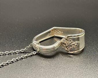 May include: A silver spoon pendant necklace. The spoon is bent into a heart shape, with floral detailing on the spoon's head. The pendant hangs from a delicate silver chain. The background is a dark, textured surface.