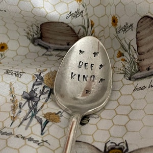 May include: A silver spoon with the words "Bee Kind" engraved on it. The spoon is lying on a white fabric with a beehive pattern.