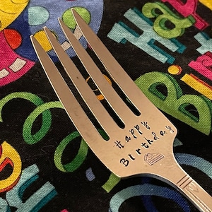 May include: A silver fork with the words "Happy Birthday" engraved on the handle. The fork is lying on a colorful fabric with a birthday theme.