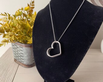 May include: A silver necklace with a large open heart pendant. The necklace is displayed on a black velvet stand.
