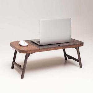 May include: A wooden laptop stand with a dark brown finish and a light brown top. The stand has two legs on each side and a white wireless mouse is on the top. A silver laptop is on the stand with the screen facing away from the camera.