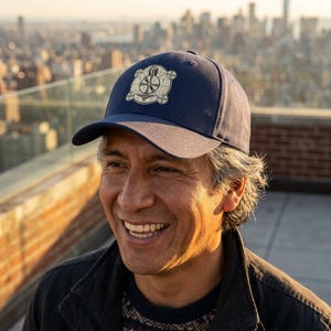 May include: A navy blue baseball cap featuring a circular graphic on the front panel. The design consists of a central wheel-like emblem surrounded by four smaller circular icons, all rendered in a light beige, distressed, vintage-style print.