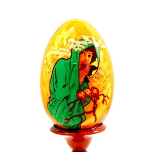 Op de afbeelding: Een handbeschilderd houten ei met een afbeelding van een vrouw in een groene mantel die een baby vasthoudt. Het ei staat op een houten standaard.