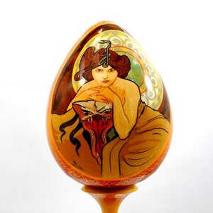 May include: A hand-painted wooden egg featuring a detailed illustration of a woman with a scorpion on her head and a mythical creature. The egg is displayed on a wooden stand.